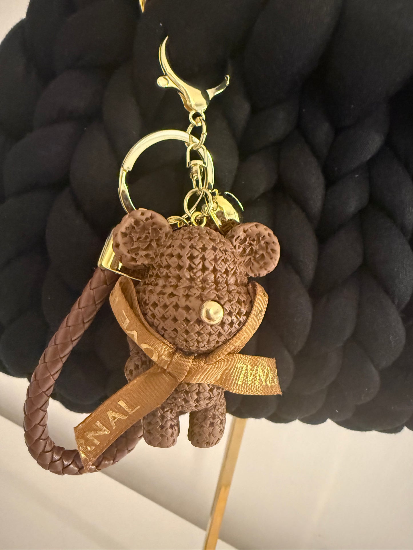 Close-up of detachable woven teddy bear charm with gold ribbon bow and braided leather loop on Nightlife black bag