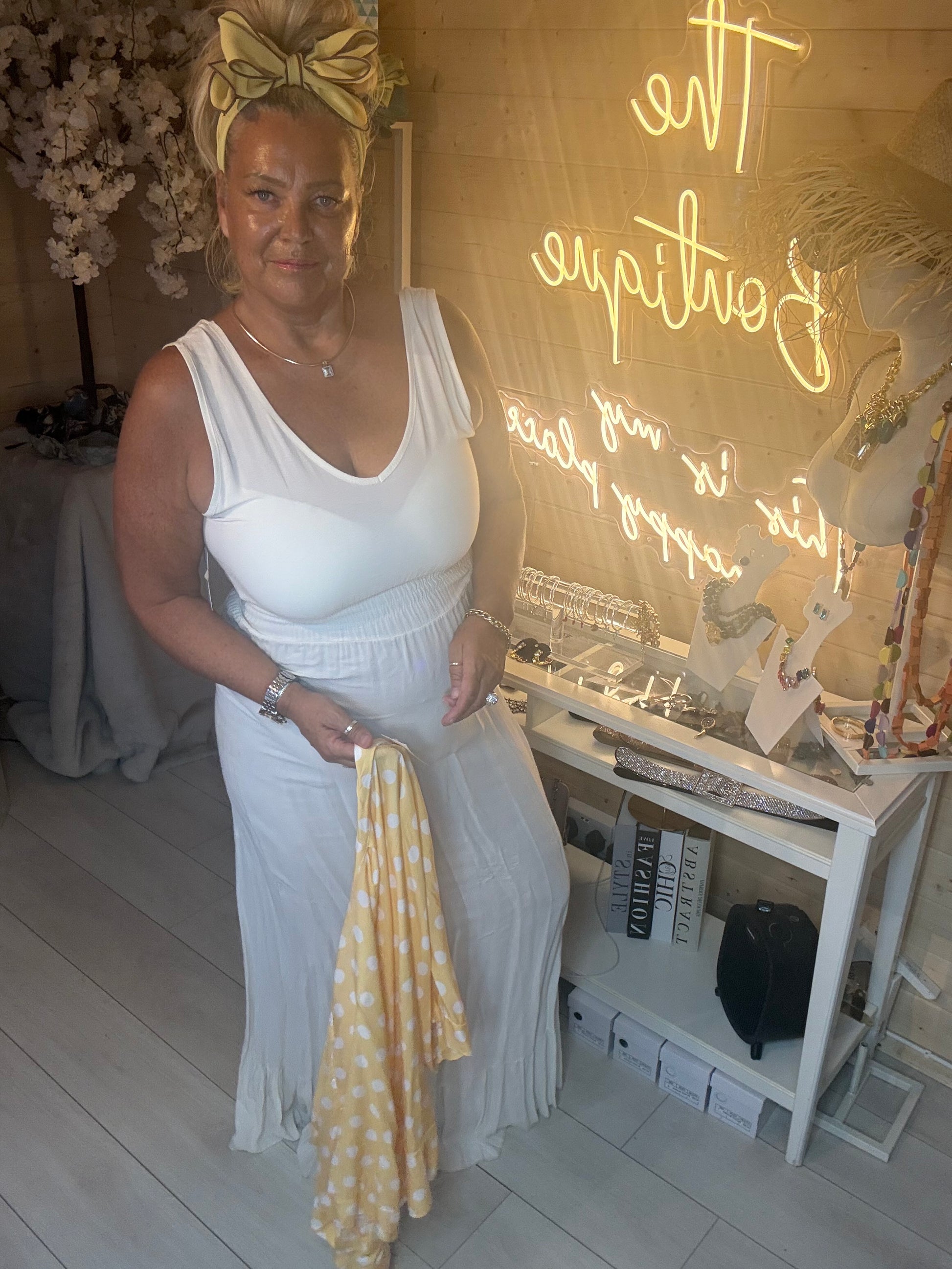 Woman wearing yellow polka dot shawl as kimono jacket over white ribbed maxi dress with white quilted bag boutique setting