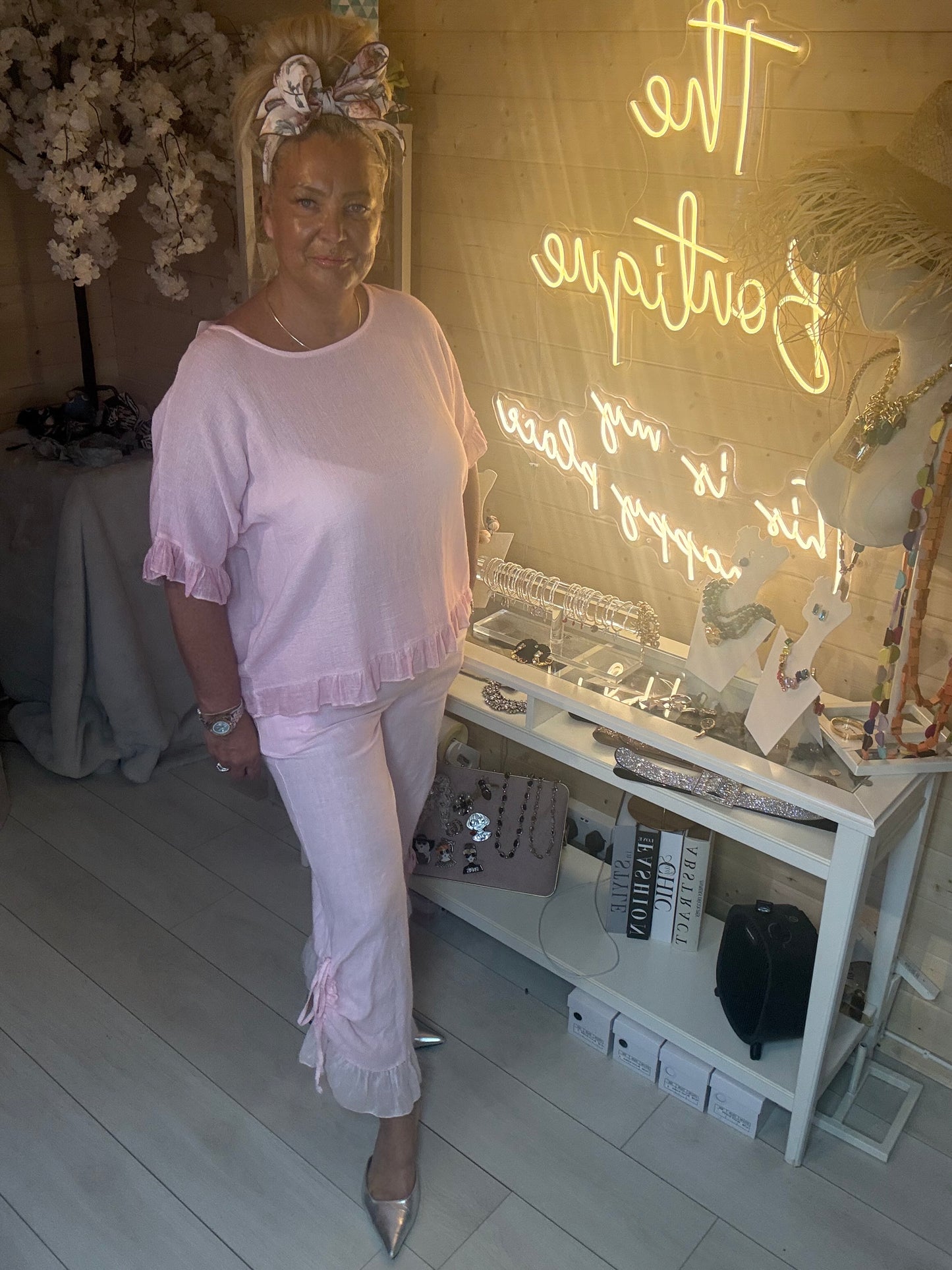 Model sitting wearing Rosa Blush Pink Ruffle Hem Crinkle Top with matching pink ruffle linen trousers boutique background