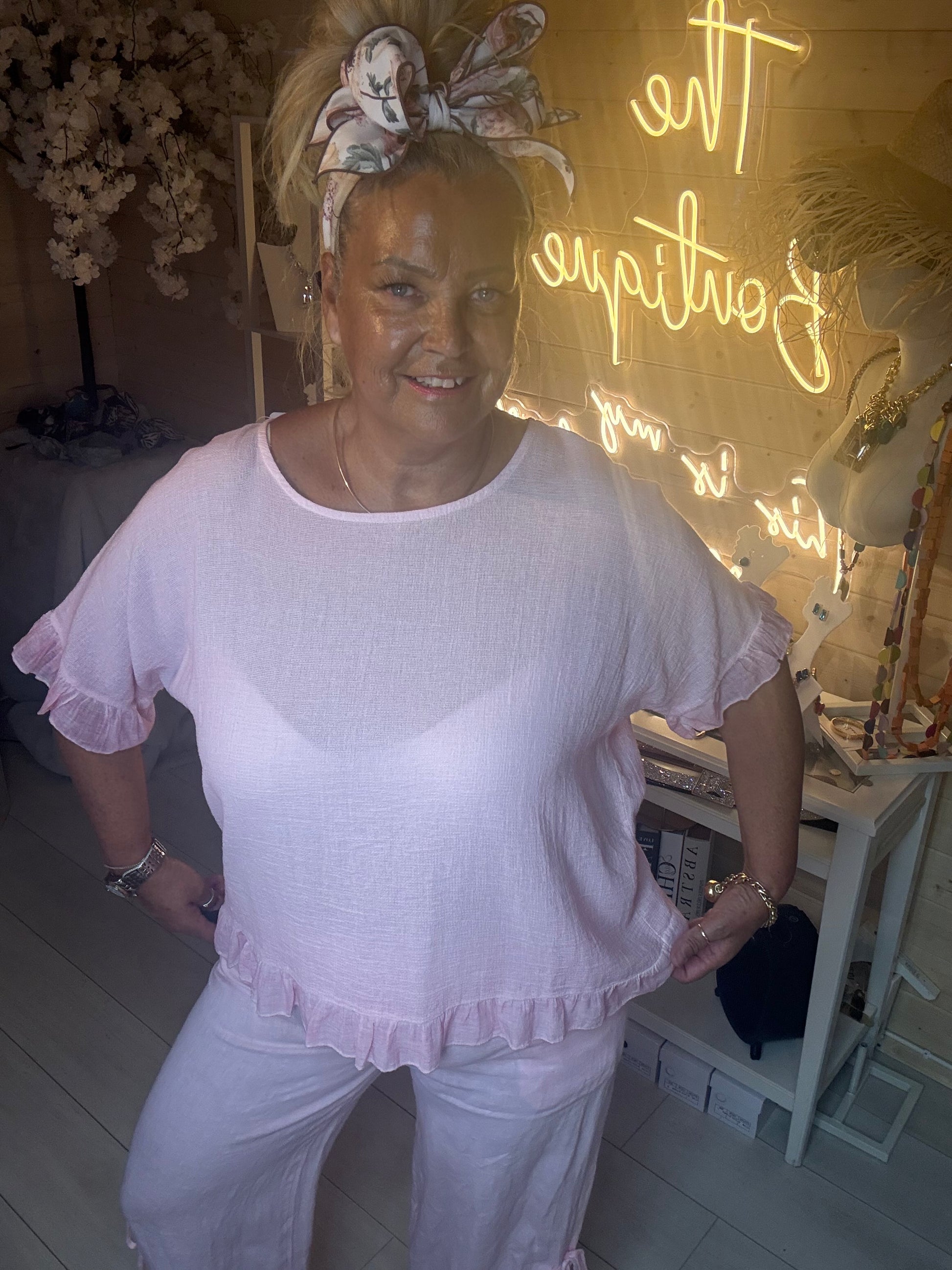 Side angle view of Rosa Blush Pink Ruffle Hem Crinkle Top with matching pink linen trousers boutique neon sign background