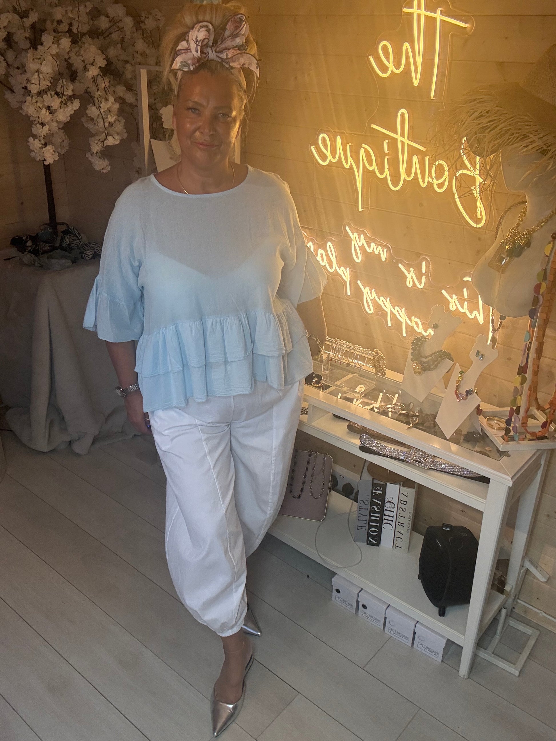 Woman wearing Sky Blue Tiered Ruffle Hem Top styled with white boom trousers and silver heels in The Ultimate Ladies Boutique