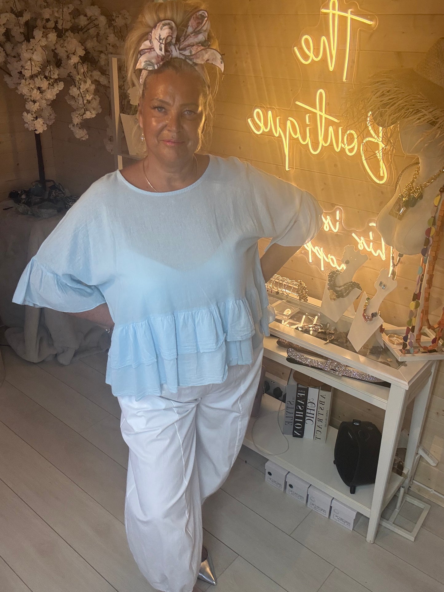 Model wearing Sky Blue Tiered Ruffle Hem Top with white boom trousers showing relaxed flattering silhouette boutique background