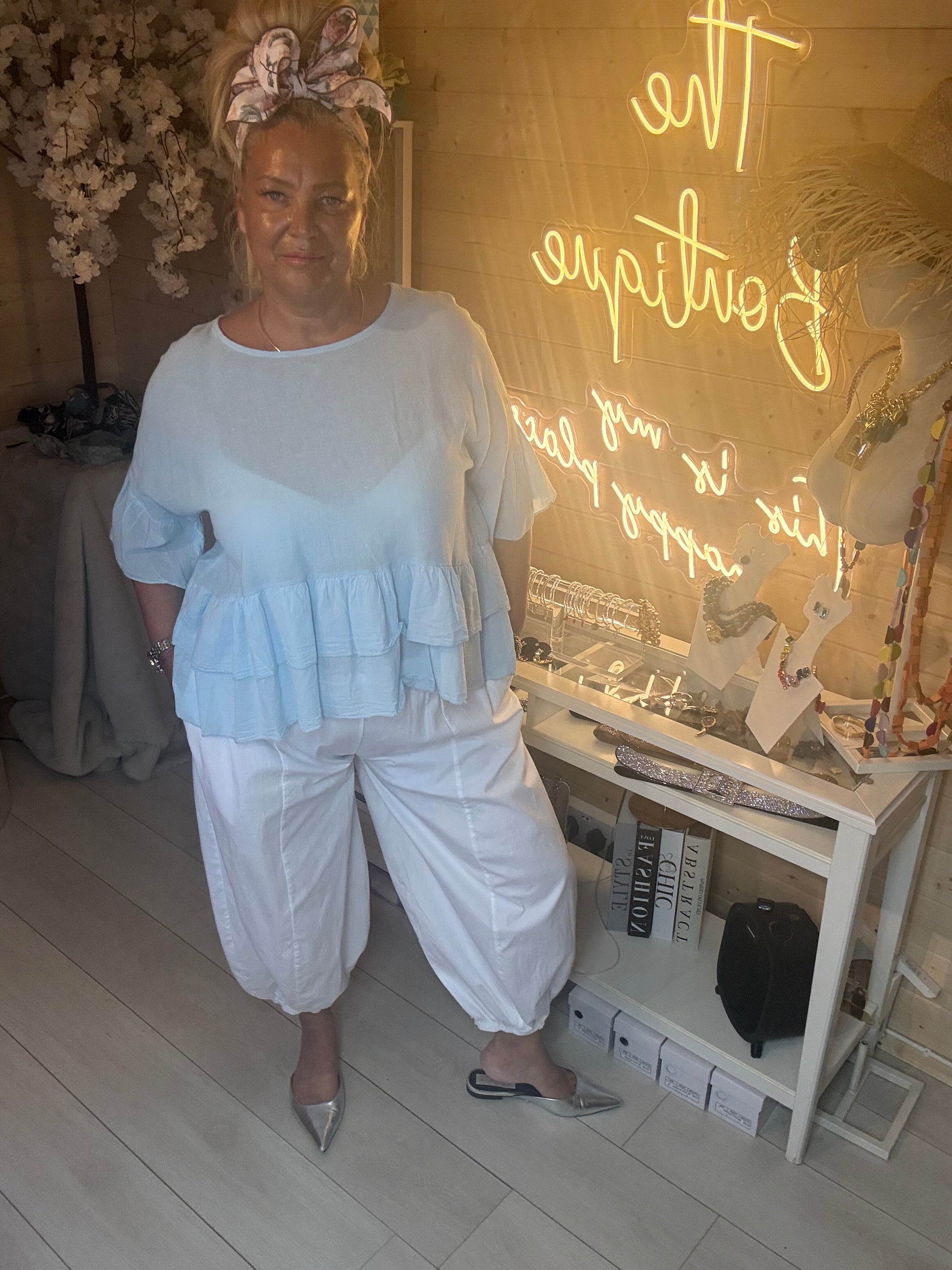 Full length view of Sky Blue Tiered Ruffle Hem Top with white balloon harem trousers and silver pointed heels boutique setting