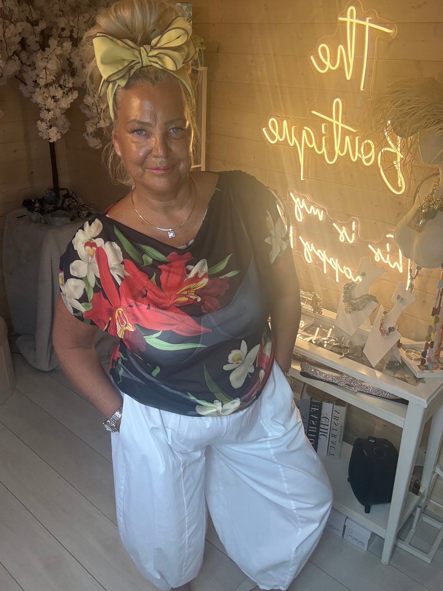 Model wearing Tropea Dark Floral Satin Top showing vibrant tropical floral print on dark navy base with white boom trousers boutique background
