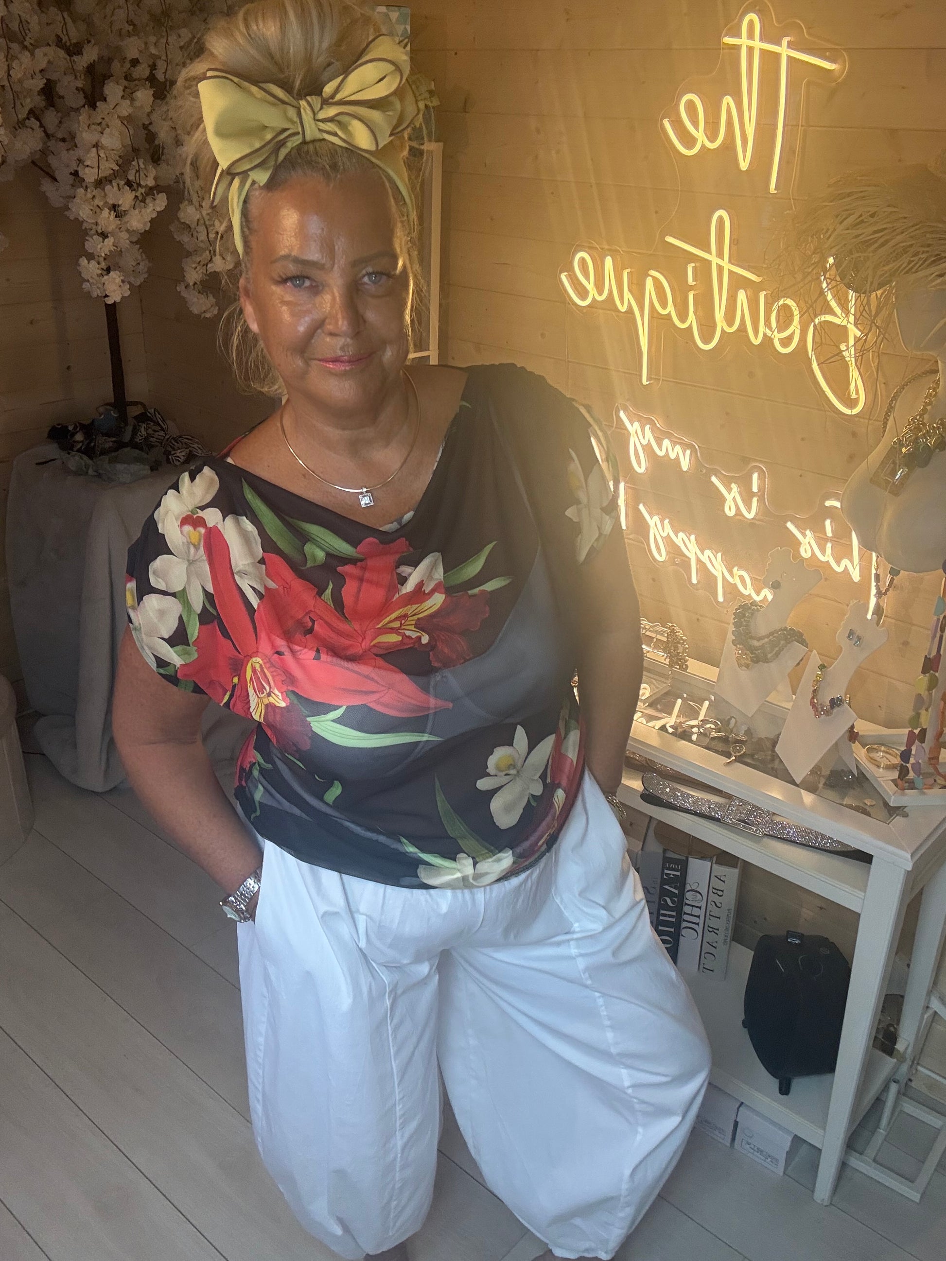 Model wearing Tropea Dark Floral Satin Top showing vibrant tropical floral print on dark navy base with white boom trousers boutique background