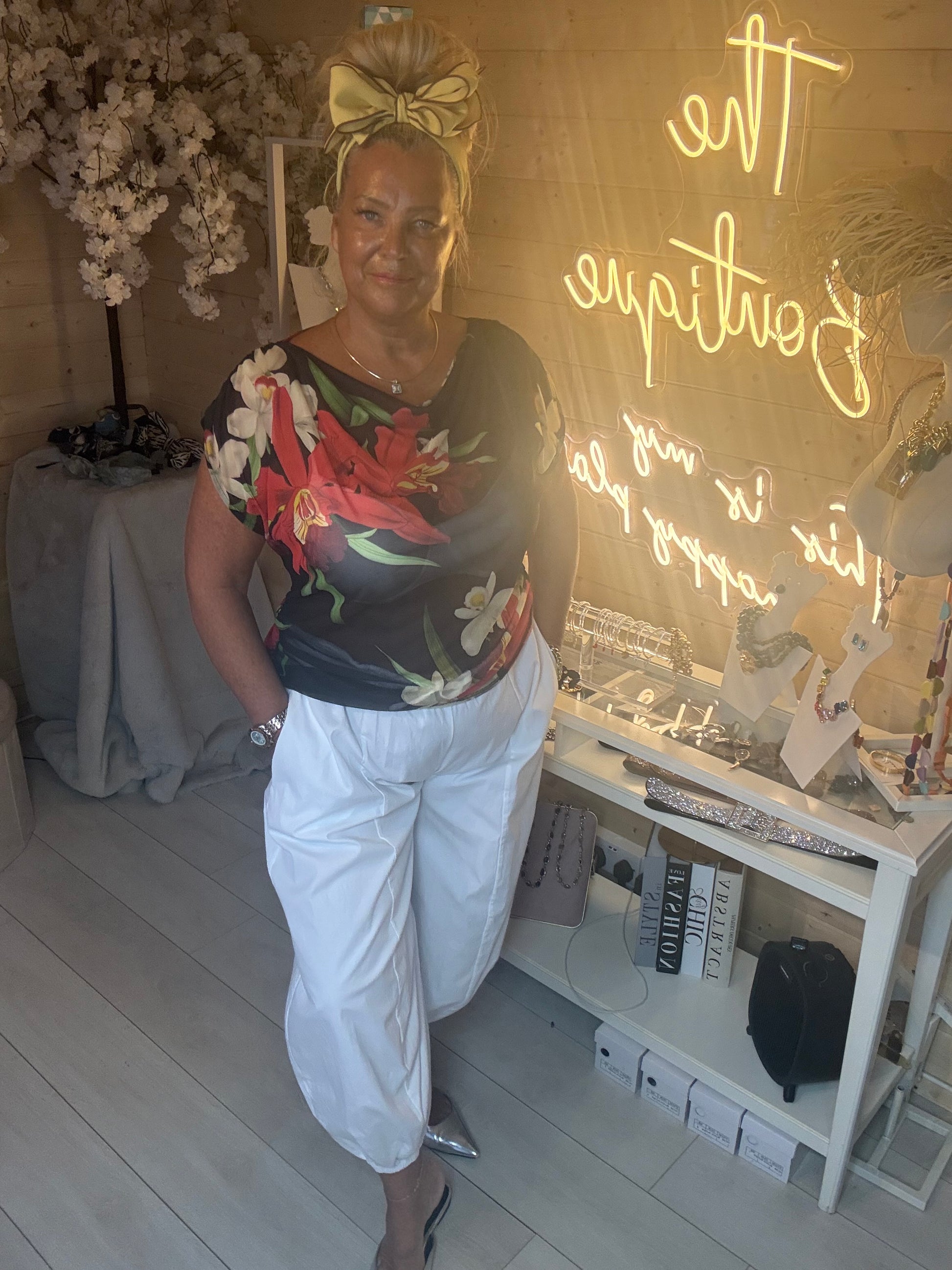 Woman wearing Tropea Dark Floral Satin Top with bold red and white tropical print styled with white boom trousers and silver heels in The Ultimate Ladies Boutique