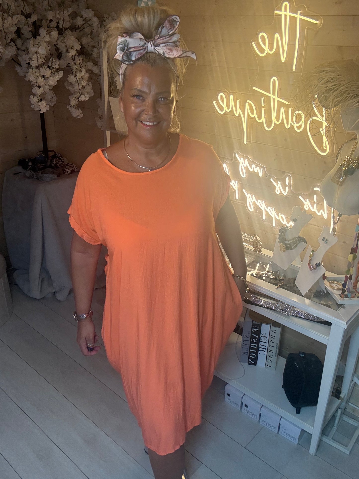 Woman wearing Coral Cocoon Twist Back Jersey Dress front view with silver heels in The Ultimate Ladies Boutique boutique neon sign background