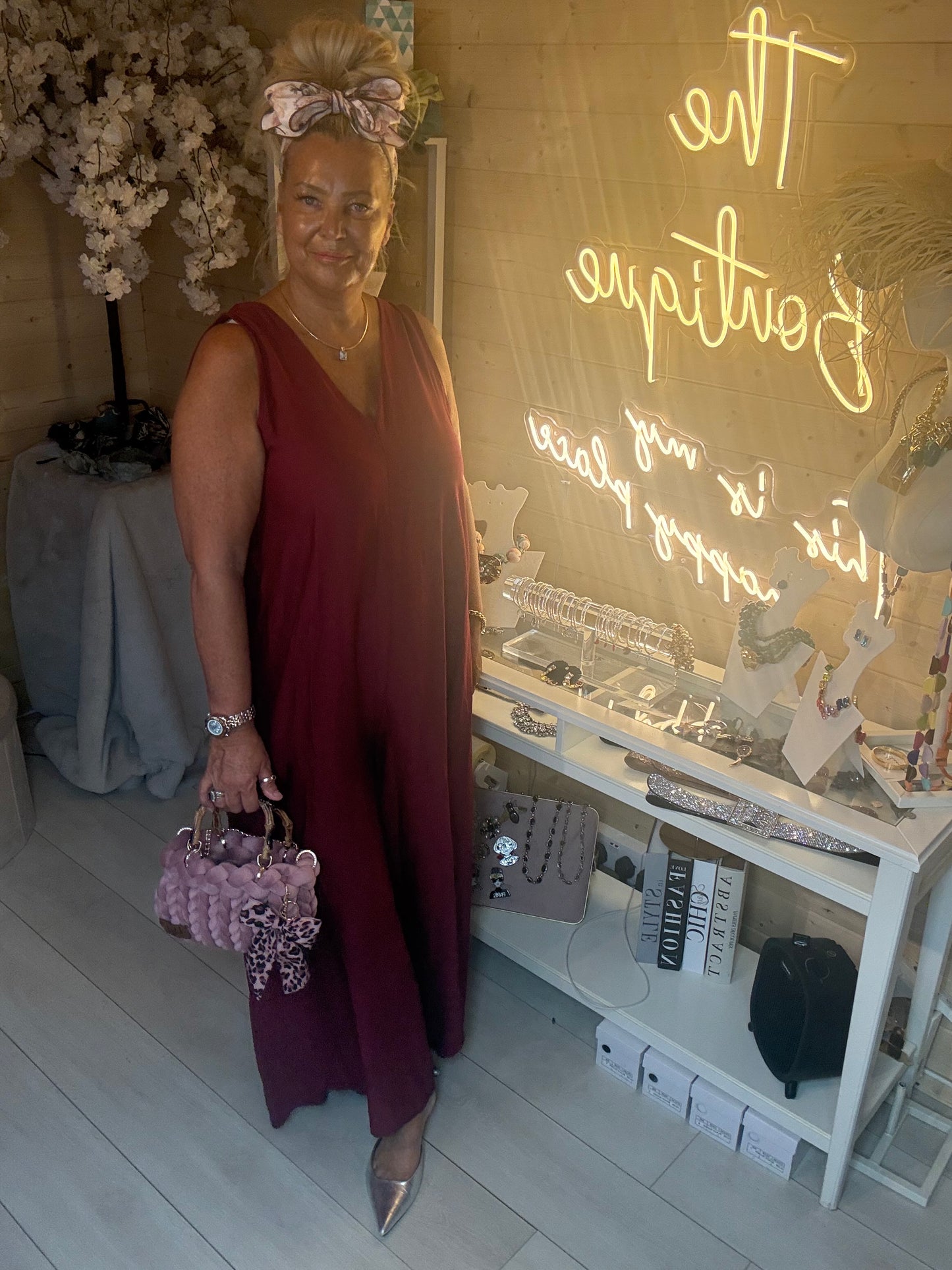 Full length view of Merlot Burgundy Sleeveless Wide Leg Jumpsuit with silver pointed heels and lilac chunky woven tote bag boutique neon sign background