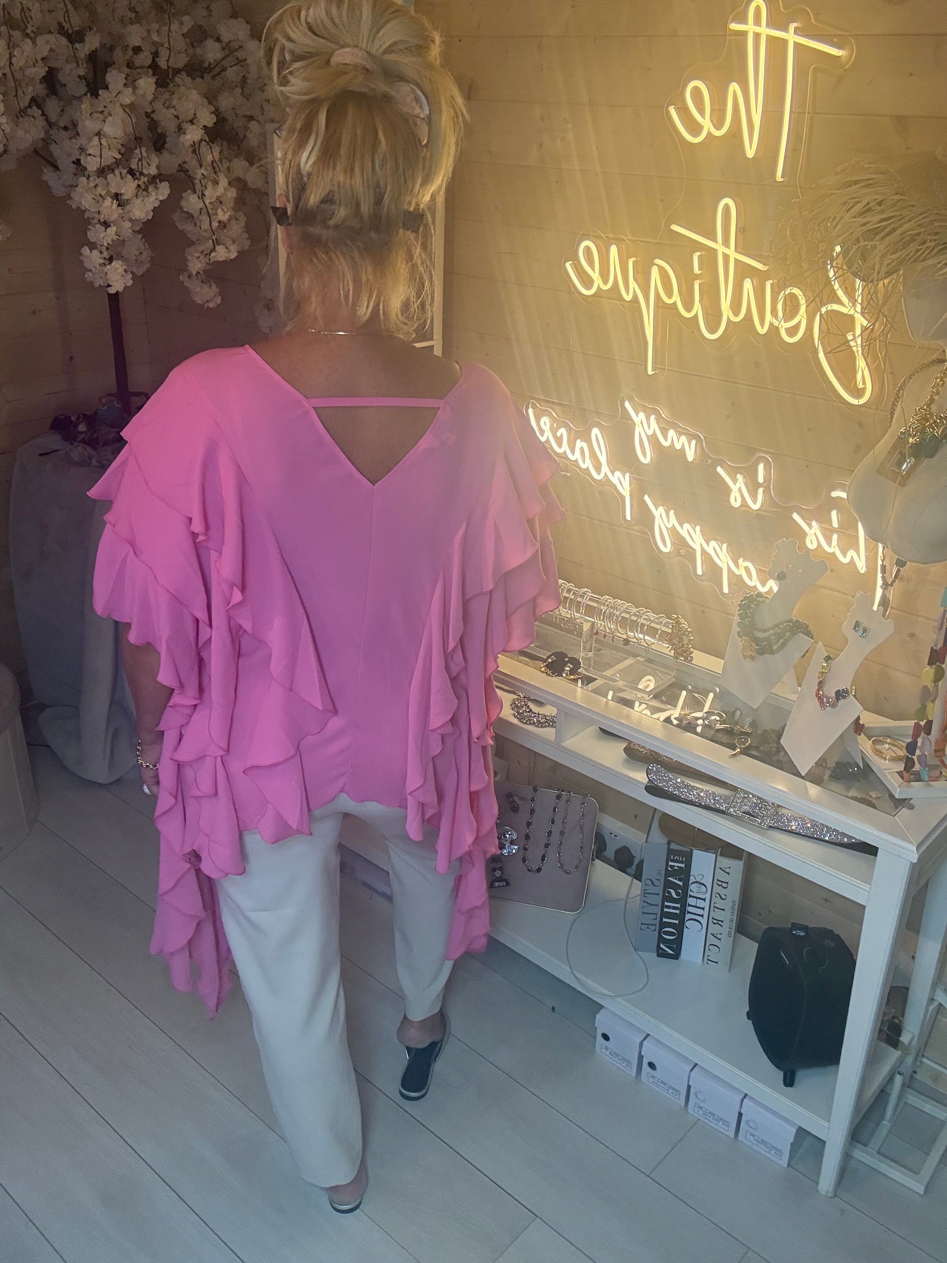 Model wearing Fiesta Hot Pink Tiered Ruffle Top showing V-neckline and tiered ruffle layers with white trousers boutique neon sign background