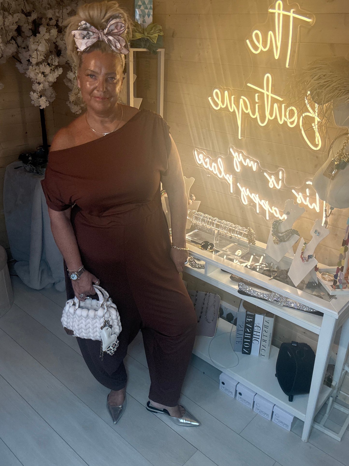 Woman wearing Coco Chocolate Brown Off-Shoulder Slouch Jumpsuit with white chunky woven mini bag and silver heels in The Ultimate Ladies Boutique