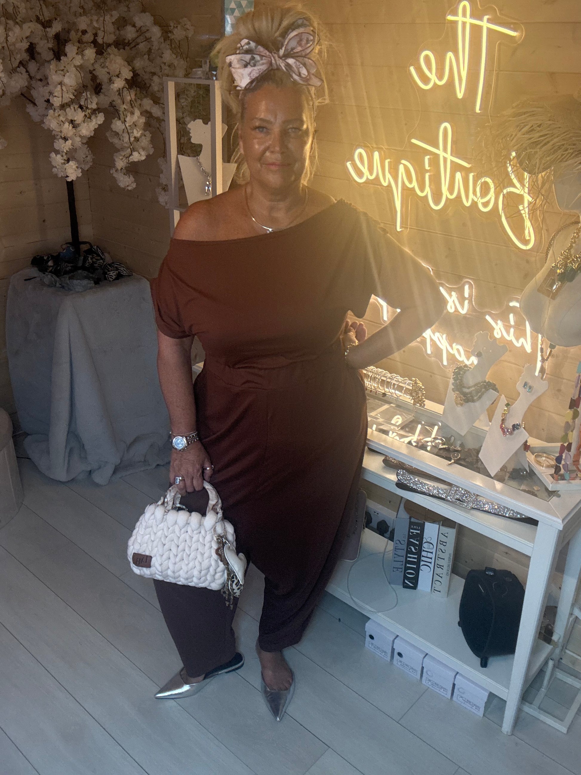 Model wearing Coco Chocolate Brown Off-Shoulder Slouch Jumpsuit showing flattering off-one-shoulder neckline with white woven bag boutique neon sign background