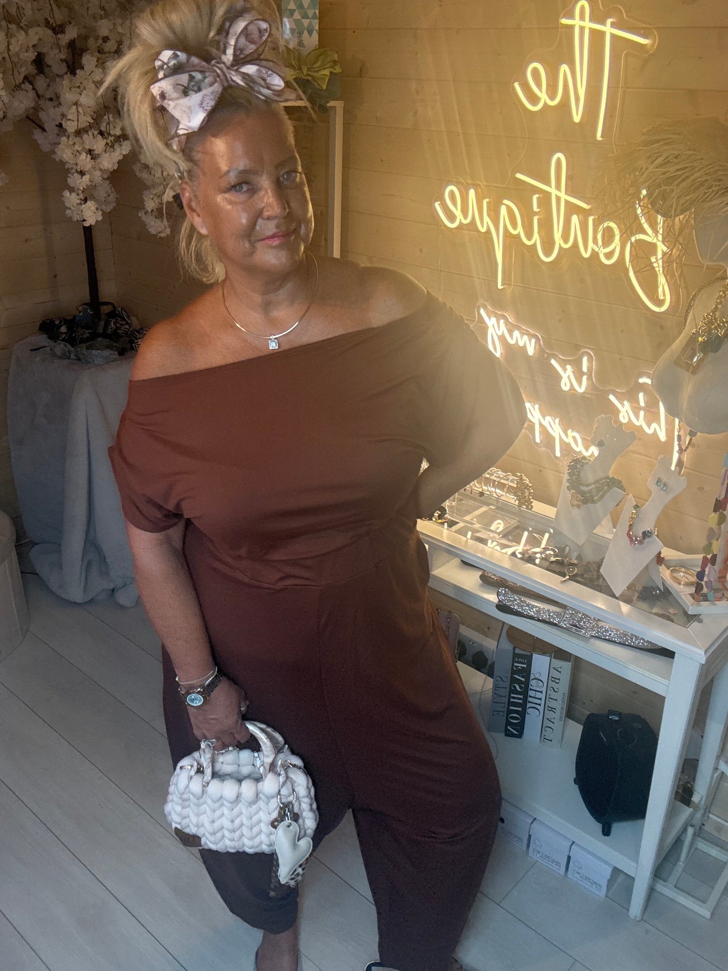 Model posing in Coco Chocolate Brown Off-Shoulder Slouch Jumpsuit showing relaxed slouchy fit and rich chocolate brown colour with white chunky woven bag one size 8-18