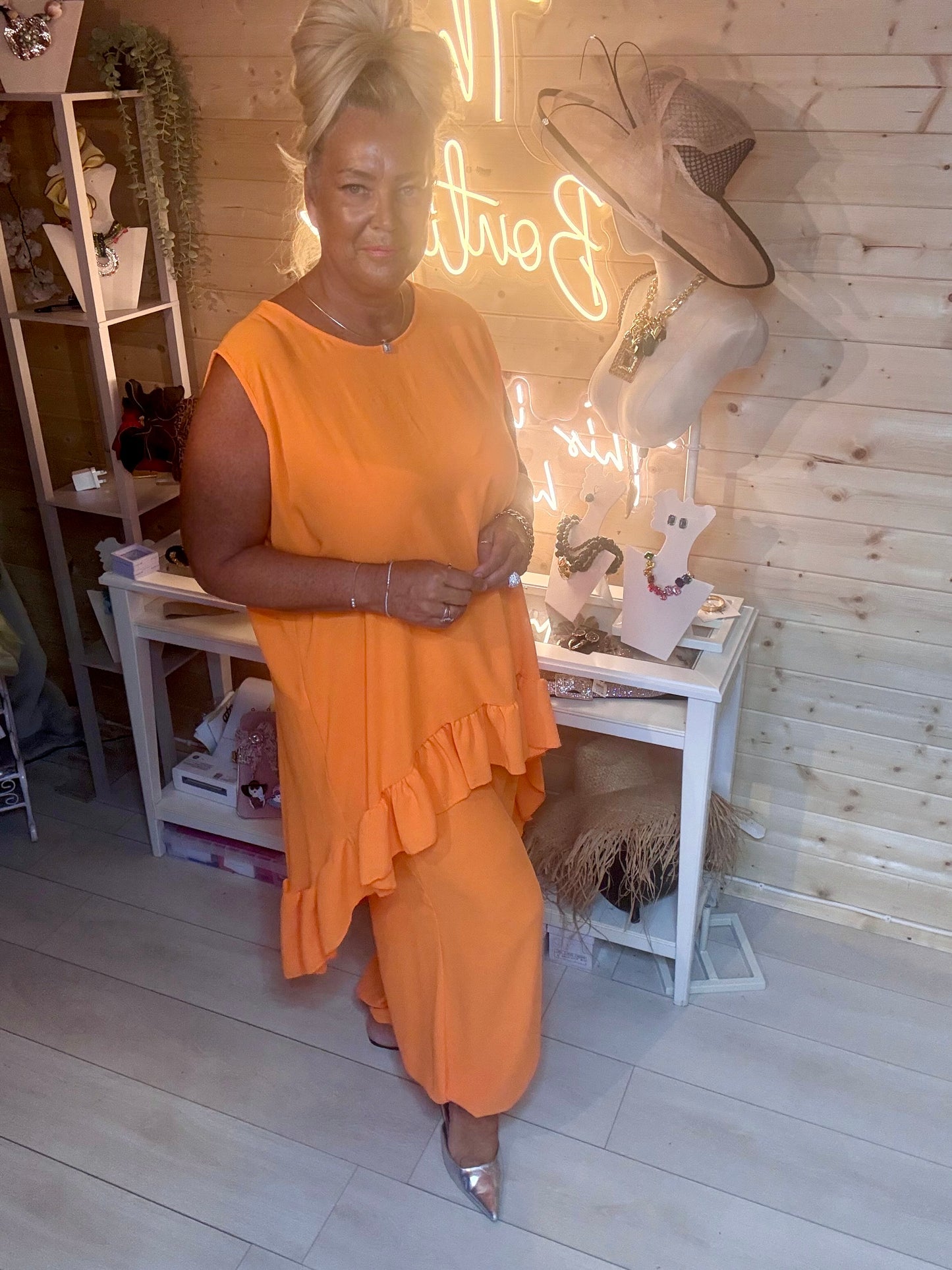 Orange ruffle hem co-ord set – full length side view showing asymmetric ruffle hem vest top and matching harem trousers with silver mules, boutique setting
