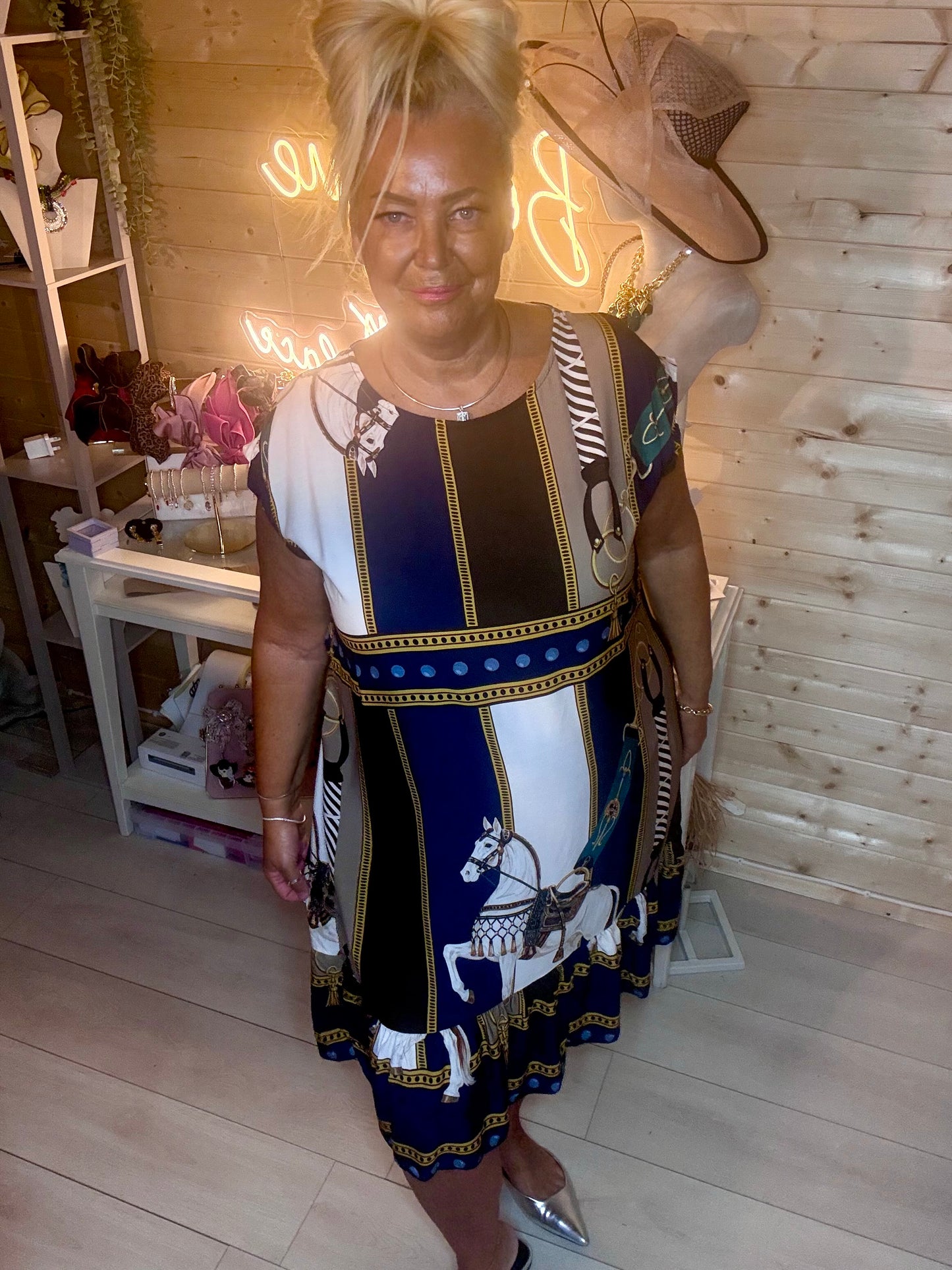 Equestrian scarf print shift dress in navy, white and gold – full length front view showing round neckline, cap sleeves, horse motif panel and tiered ruffle hem styled with silver mules in boutique setting