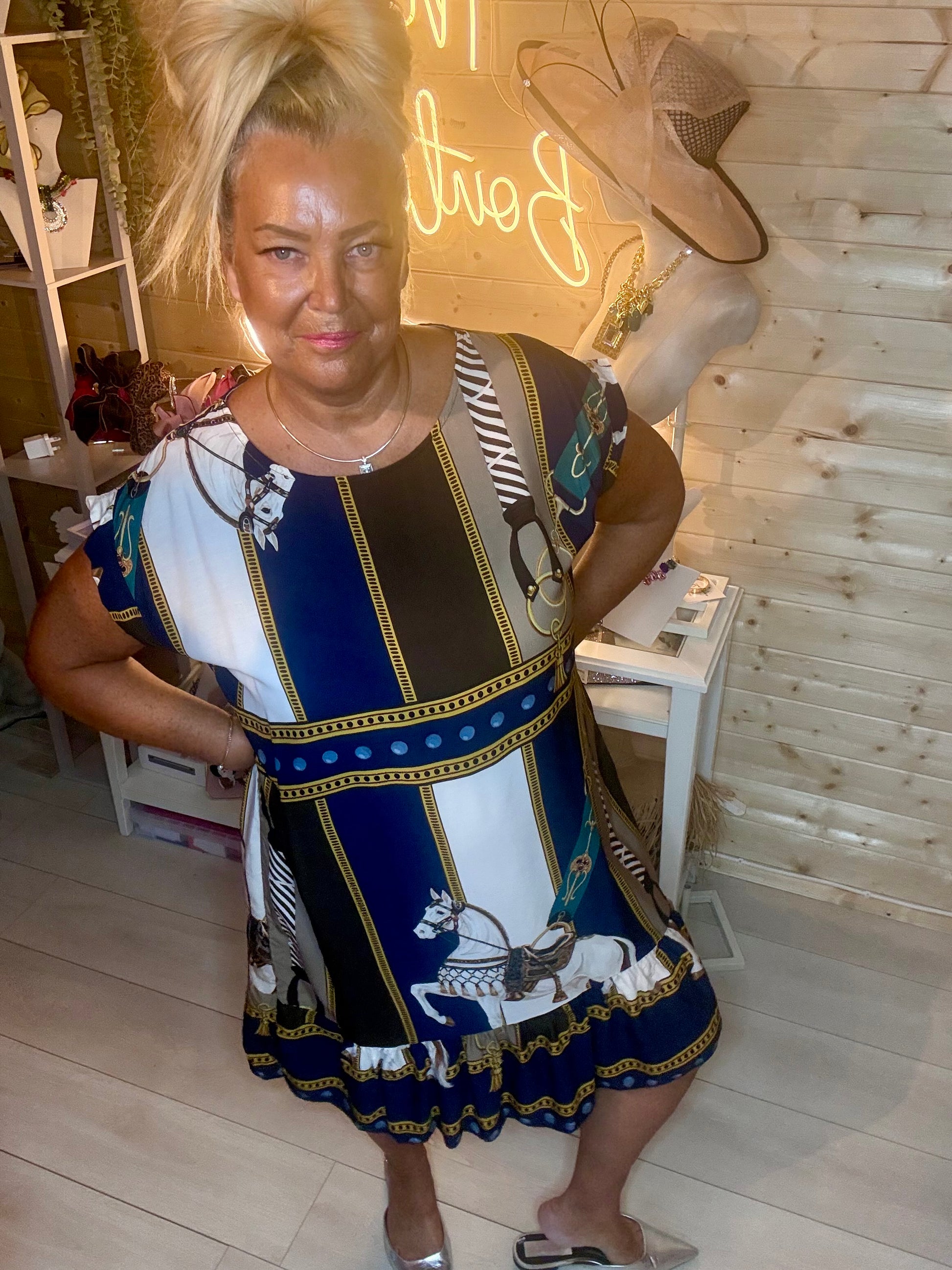 Equestrian scarf print shift dress – full length side view showing relaxed silhouette, cap sleeves and tiered ruffle hem in navy, white and gold horse print in boutique setting