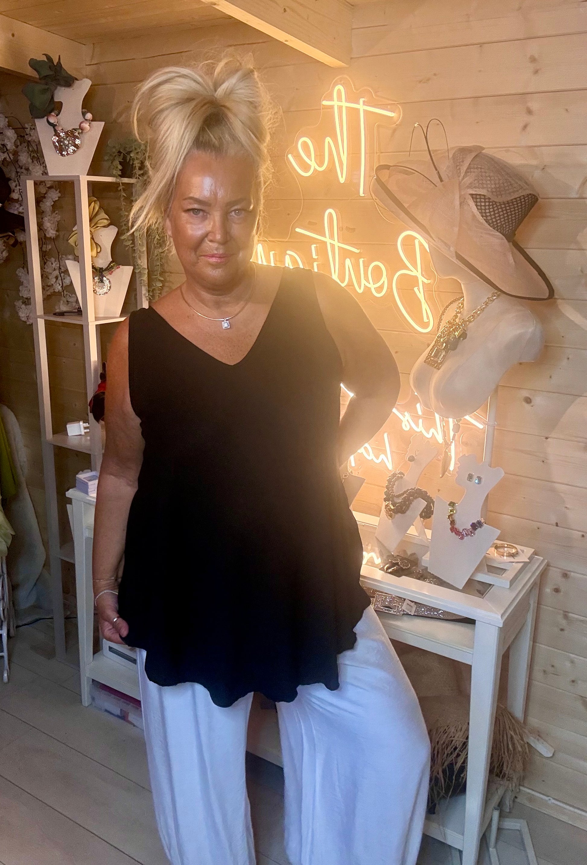 Black V-neck swing vest top – full length front view showing deep V-neckline, sleeveless design and curved hem styled with white wide-leg trousers and silver mules