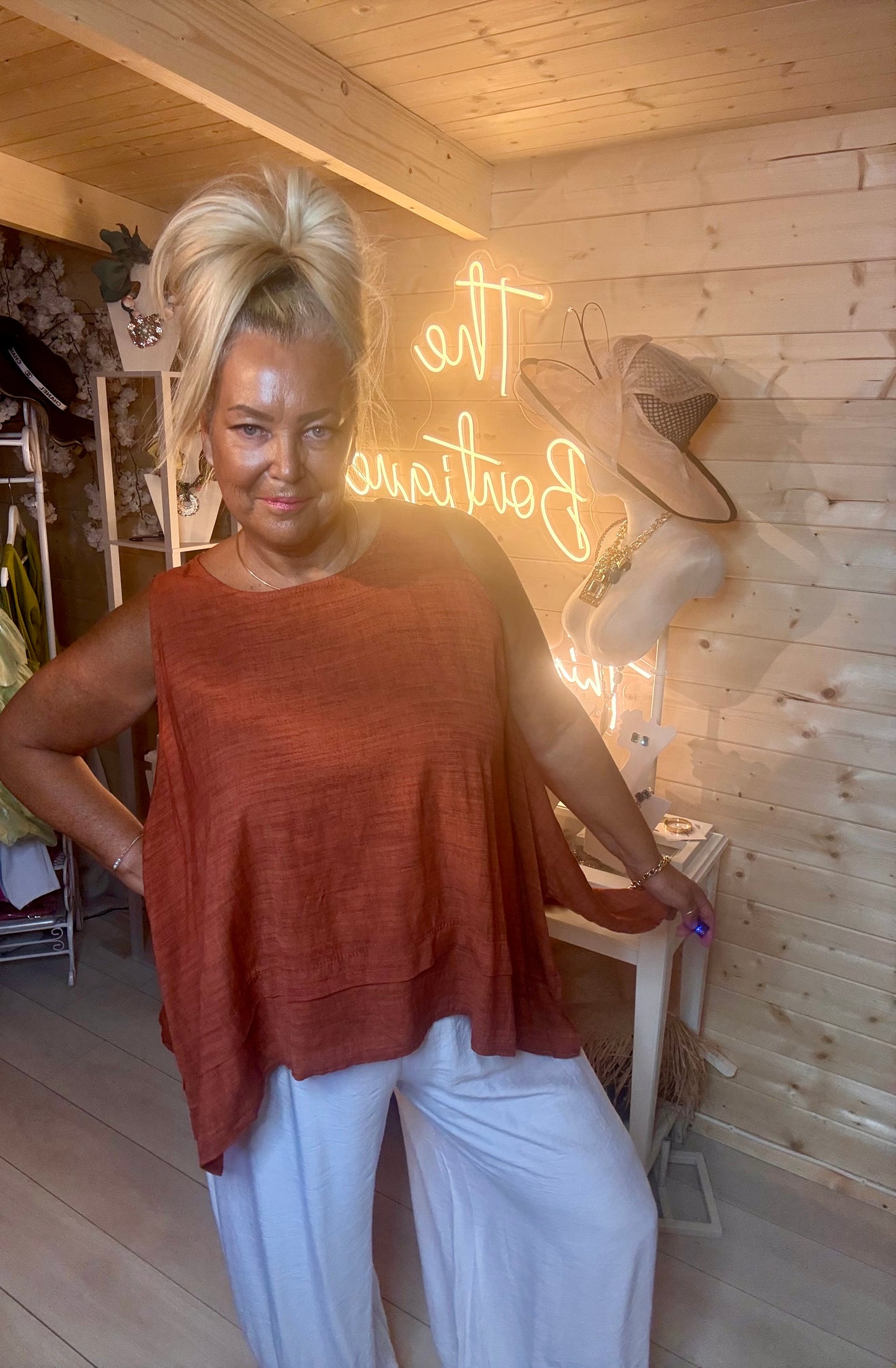 Rust terracotta fine knit swing top – full length front view showing round neckline, short sleeves and layered double hem styled with white wide-leg trousers and silver mules in boutique setting