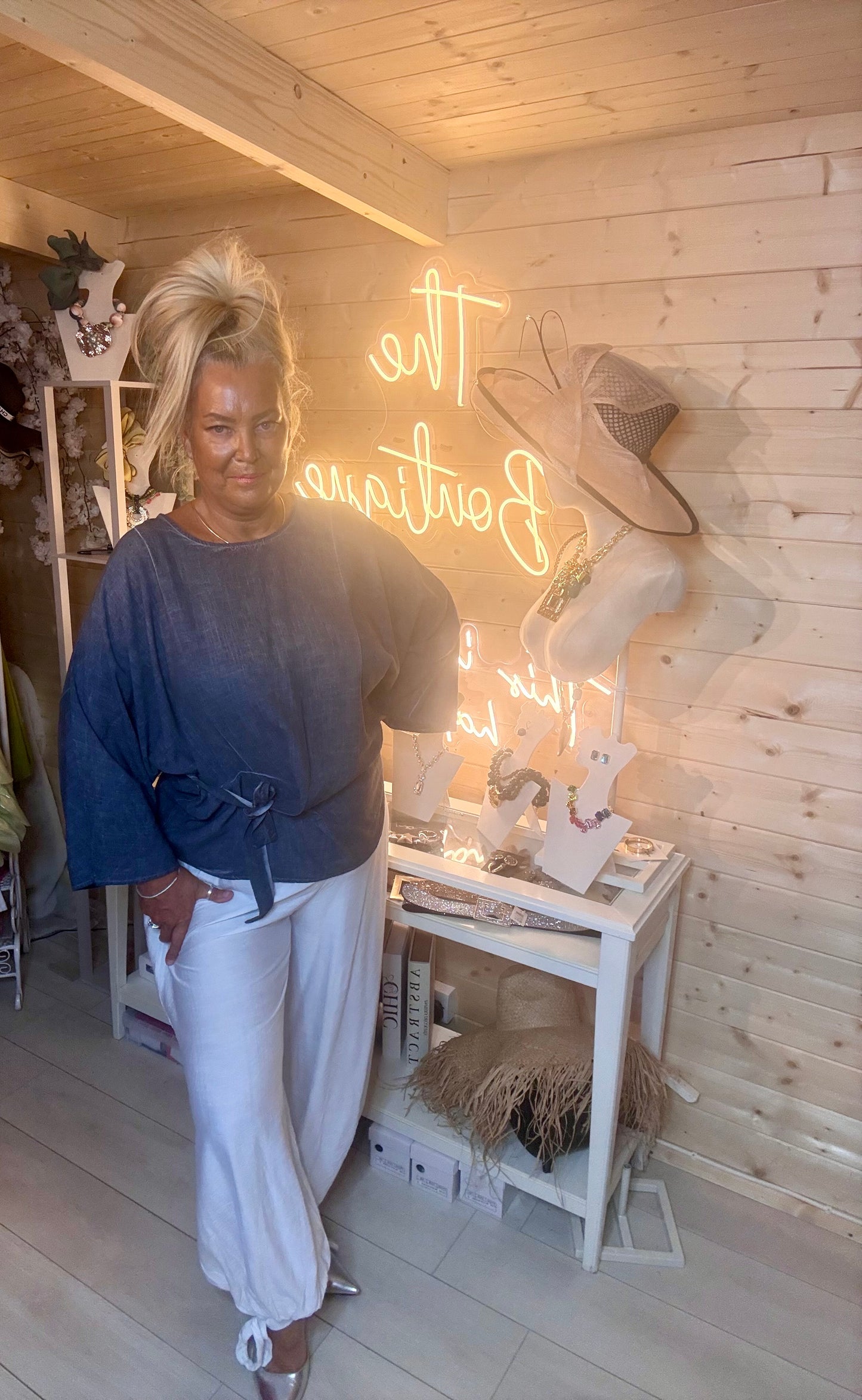 Charcoal grey wrap tie batwing top – full length front view showing round neckline, batwing sleeves and waist-cinching wrap tie detail styled with white wide-leg trousers and silver mules in boutique setting