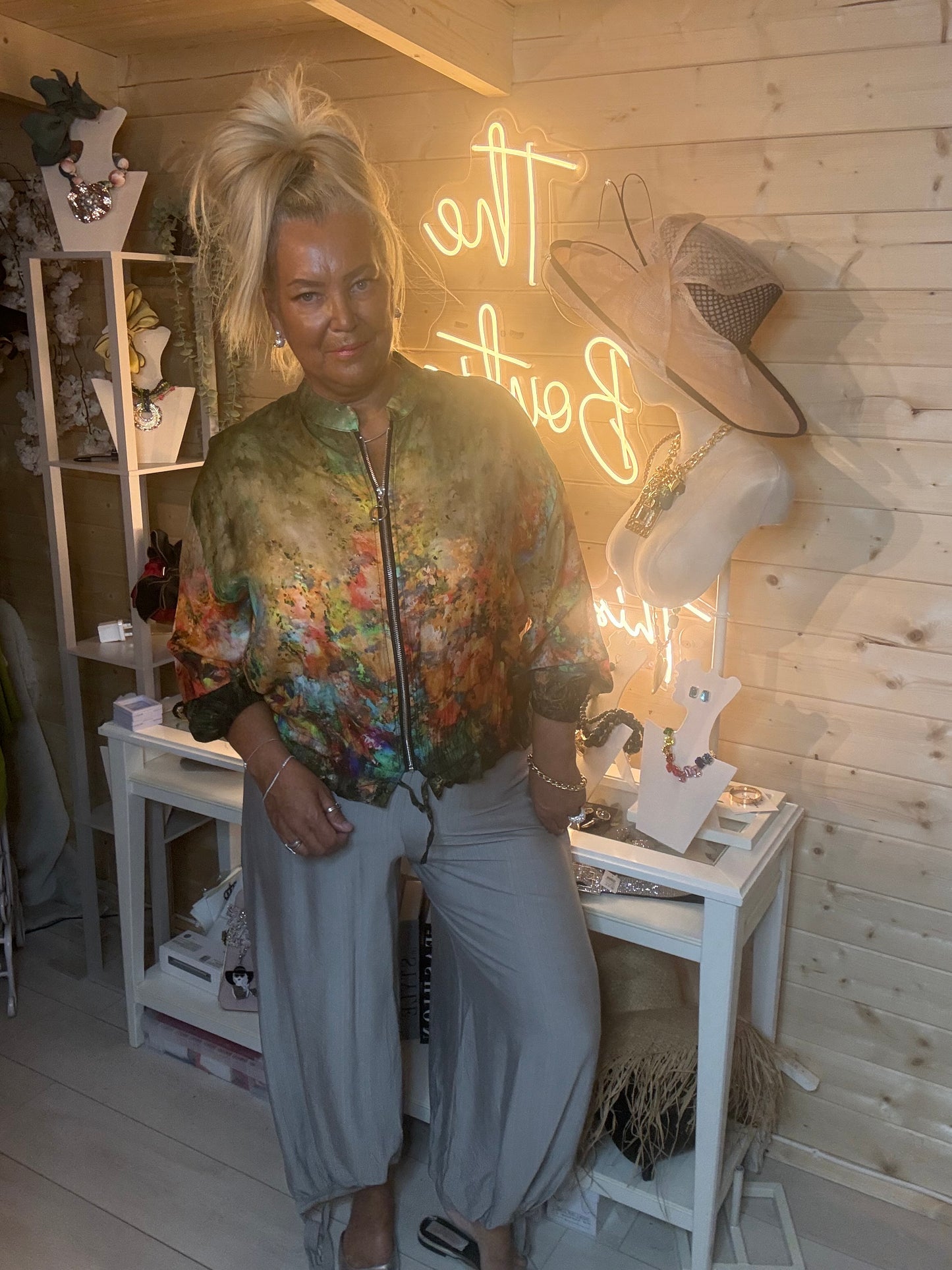 Floral print zip-up bomber jacket in rich green, orange and purple painterly print – full length front view showing zip fastening and batwing sleeves styled with grey wide-leg trousers in boutique setting
