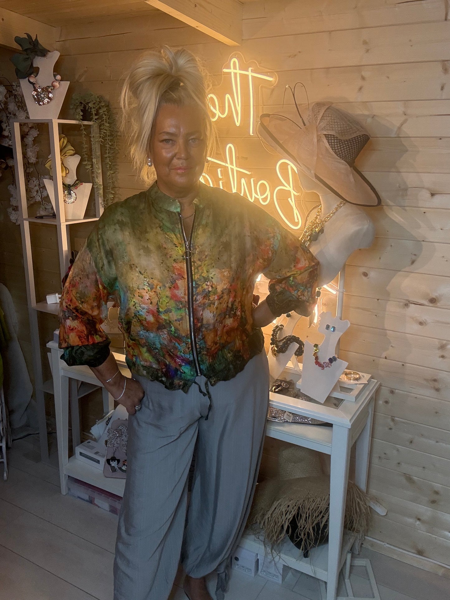 Floral print sheer bomber jacket – full length side view showing zip front, batwing sleeves and elasticated cuffs in rich green and orange floral print with grey trousers